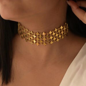 May include: A gold-colored choker necklace with a repeating star pattern. The necklace is worn around the neck and is made of multiple rows of interconnected star shapes. The background is a blurred view of a person wearing the necklace.