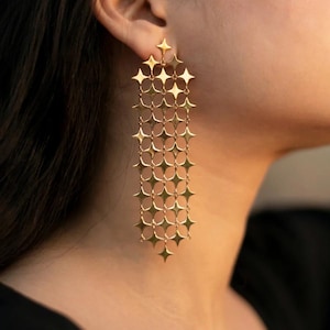 May include: Gold-toned, long dangle earrings. Each earring features a star-shaped top, with multiple star-shaped links cascading downwards. The earrings are designed to be worn, and are shown against a blurred background.