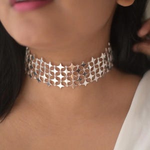 May include: A silver choker necklace with a repeating starburst pattern. The necklace is made of interconnected, flat metal pieces, creating a wide band that sits around the neck. The design is intricate and geometric, reflecting light.
