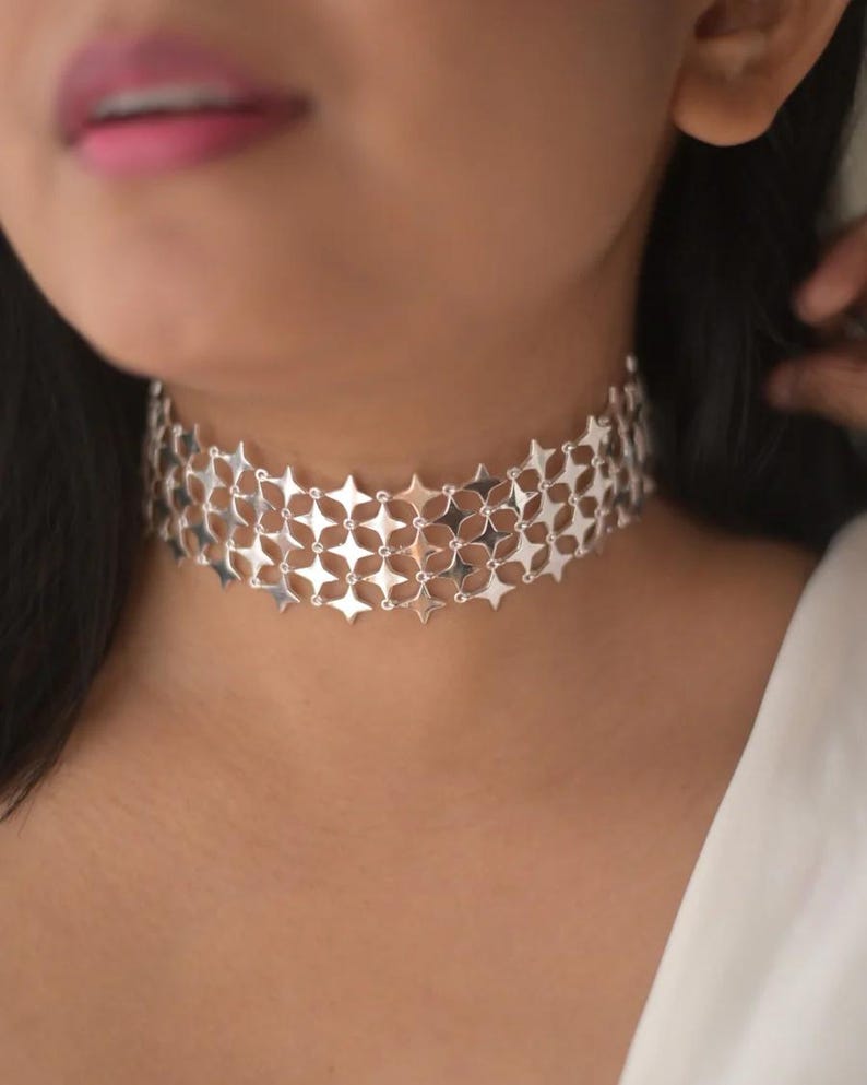 May include: A silver choker necklace with a repeating starburst pattern. The necklace is made of interconnected, flat metal pieces, creating a wide band that sits around the neck. The design is intricate and geometric, reflecting light.