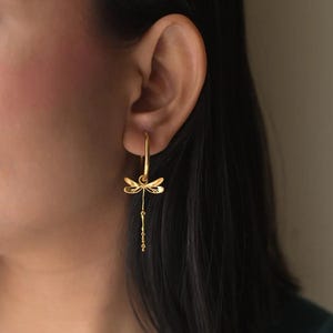 Può includere: Orecchini dorati a forma di libellula. Gli orecchini presentano un design a libellula con ali delicate e una catena pendente di piccole perle. L'attenzione è focalizzata sui gioielli.
