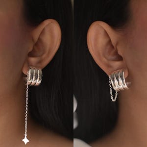 May include: Silver earrings with a stacked, curved design that sits on the earlobe. A fine chain hangs from the earrings, terminating in a small, four-pointed star. The earrings are displayed on a person with dark hair.