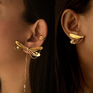 Può includere: Orecchini a forma di libellula color oro con dettagli intricati delle ali e catene pendenti. Un orecchino presenta una goccia di perla. L'altro orecchino ha una piccola gemma. È visibile anche un ear cuff dorato.