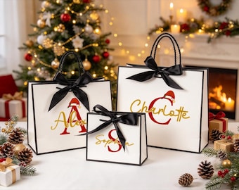 Pode incluir: Três sacos de presente brancos com alças de corda preta e laços de cetim preto. Cada saco apresenta um nome personalizado em vermelho e dourado com um pequeno gráfico de gorro de Pai Natal na primeira letra: Alex, Sophia e Charlotte.