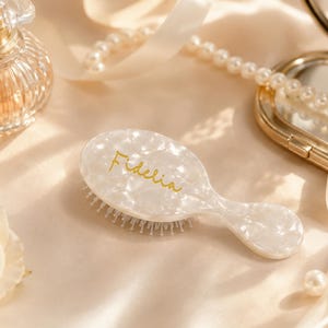 May include: A small, oval-shaped hairbrush with a white, pearlescent, marbled finish. The name Fidelia is written in gold cursive script on the center of the brush handle. The brush features white bristles and a smooth, glossy, reflective surface.