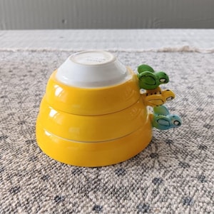 May include: A stack of yellow ceramic measuring cups with a white top. The cups are adorned with three colorful frog handles in green, yellow, and blue. The cups are sitting on a patterned surface.