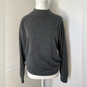 May include: A dark gray mock neck sweater with long sleeves. The sweater is displayed on a mannequin. The fabric appears to be a soft knit. The sweater has a ribbed neckline and cuffs.