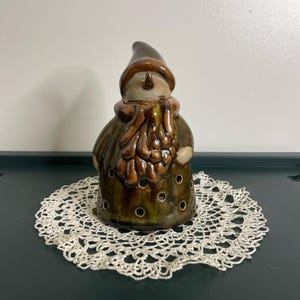 May include: A ceramic snowman ornament with a brown hat and scarf, and a green coat. The snowman has a beige face and a carrot nose. The base has circular openings. The ornament sits on a white crocheted doily. A festive decoration.