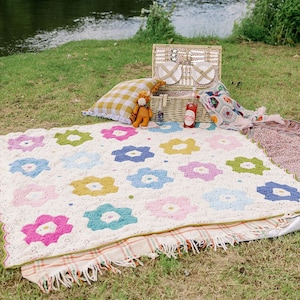 May include: A picnic setting featuring a cream-coloured blanket adorned with crochet flower patterns in various colours. A wicker picnic basket, a yellow gingham cushion, and a soft toy are visible. A river and green grass are in the background.