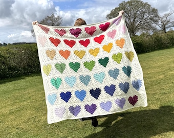 May include: A white crochet blanket with a rainbow of hearts. The blanket is made with a granny square stitch and features a variety of colors, including red, orange, yellow, green, blue, indigo, and violet.