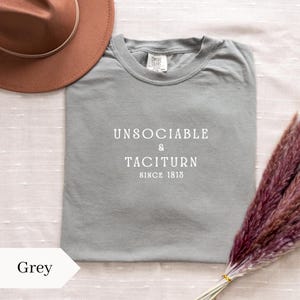 Puede incluir: Una camiseta gris Comfort Colors con las palabras "UNSOCIABLE & TACITURN SINCE 1813" impresas en blanco. Un sombrero marrón y un ramo de flores moradas secas también están en la imagen.