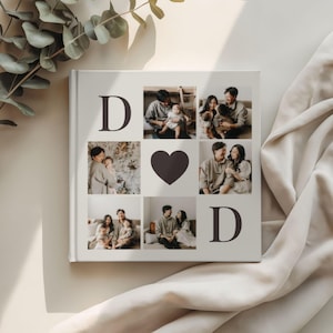 Puede incluir: Un álbum de fotos cuadrado de color blanco roto con la letra "D" en las esquinas superior izquierda e inferior derecha. El álbum presenta fotos familiares dispuestas en una cuadrícula, con un corazón oscuro en el centro. Las fotos muestran una familia.