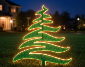 Op de afbeelding: Een groene, verlichte kerstboom tuindecoratie. De boom heeft een glittertextuur en is omrand met een warm, gloeiend licht. Hij staat in een grasrijke tuin in de schemering, met een huis en een straatlantaarn op de achtergrond.
