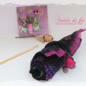 May include: A rolled up piece of fabric with a black background and purple and pink accents. The fabric is rolled up and tied with a black ribbon. A wooden crochet hook is lying on a white surface next to the fabric.
