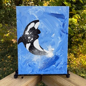 May include: Acrylic painting of a black and white orca leaping out of blue water. The background is a textured blue with white accents, suggesting waves and splashes. The artwork is displayed on a small black easel.