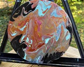 Puede incluir: Pintura abstracta redonda con un diseño de arte fluido. La obra presenta una mezcla de colores, incluyendo naranja, azul, rosa y negro, creando un efecto marmolado. La pintura se exhibe en un caballete negro, mostrando la paleta de colores única.