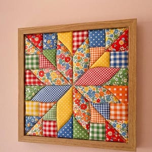 May include: A square wall art piece featuring a colorful fabric patchwork design in a light wood frame. The quilt-like pattern consists of various geometric shapes in red, blue, yellow, green, and orange, showcasing floral, gingham, and polka dot prints.