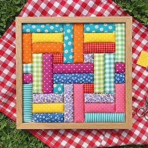 Puede incluir: Un marco de madera cuadrado que contiene un mosaico de bloques rectangulares cubiertos de tela en varios colores y patrones, incluyendo amarillo, naranja, rosa, azul y verde con estampados florales, lunares, cuadros y rayas.