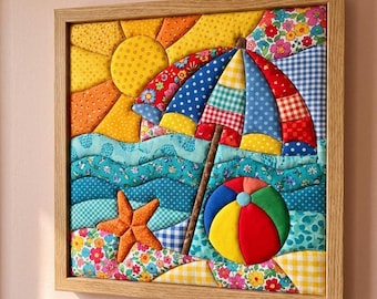 May include: A square fabric wall art piece in a light wood frame featuring a quilted beach scene. The design includes a yellow sun, a multi-coloured patterned beach umbrella, blue wavy ocean fabric, an orange starfish, and a round beach ball.