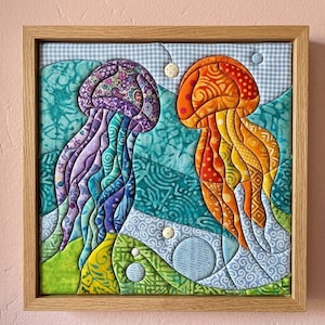 May include: A square fabric wall art piece in a light wood frame featuring two quilted jellyfish. One jellyfish is purple and violet, the other is orange and yellow. The background consists of blue and green patterned fabric patches with small circular accents.