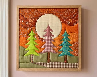 May include: A square fabric wall art piece in a light wood frame featuring a quilted landscape. The scene includes three stylised fabric trees in lime green, mauve gingham, and teal floral patterns set against a background of orange paisley and floral fabric patches with a large, circular cream-coloured moon.