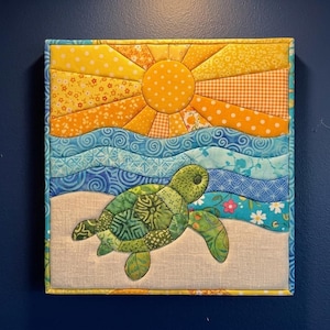May include: A square fabric wall hanging featuring a quilted scene of a green sea turtle on a beige sand beach. Above the turtle are wavy blue fabric strips representing ocean waves, topped by a yellow and orange sun with radiating rays.