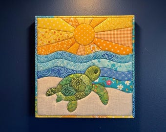 May include: A square fabric wall hanging featuring a quilted scene of a green sea turtle on a beige sand beach. Above the turtle are wavy blue fabric strips representing ocean waves, topped by a yellow and orange sun with radiating rays.