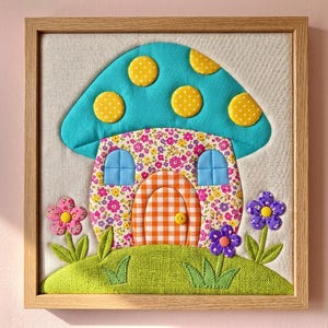 Può includere: Un'opera d'arte murale in tessuto incorniciata raffigurante una casa a forma di fungo con un cappello color ottanio decorato con pois gialli. La casa ha un corpo a motivi floreali, una porta a quadretti arancione con un pomello a bottone giallo e due finestre blu. Due fiori in tessuto poggiano su una base d'erba verde.
