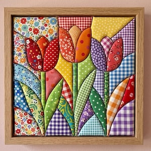 May include: A square fabric wall art piece featuring three quilted tulips in red, orange, and purple, set against a background of various colorful patterned textiles including gingham, floral, and polka dot prints, all encased in a light wood frame.