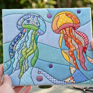 May include: A square fabric wall hanging featuring a quilted applique design of two jellyfish. The left jellyfish is blue and green with floral patterns, while the right one is yellow and orange with checkered and polka dot fabric patches.