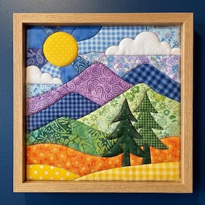 May include: A square fabric wall art piece in a light wood frame featuring a quilted landscape scene. The design includes a yellow sun, white clouds, purple and green mountains, and two dark green pine trees over orange and yellow foregrounds.