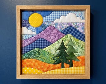 May include: A square fabric wall art piece in a light wood frame featuring a quilted landscape scene. The design includes a yellow sun, white clouds, purple and green mountains, and two dark green pine trees over orange and yellow foregrounds.