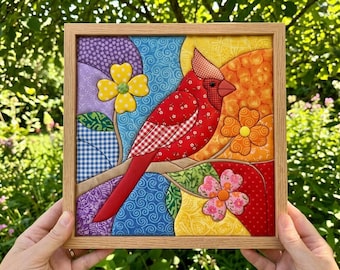 May include: A square fabric wall art piece in a light wood frame featuring a red cardinal bird perched on a branch. The design uses various patterned fabrics in red, yellow, orange, blue, and purple, with three dimensional fabric flowers.