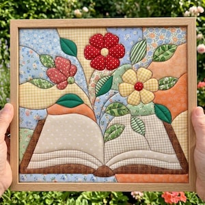 Op de afbeelding: Een ingelijst stoffen wandkunstwerk met een doorgestikt ontwerp van een open boek waar bloemen en bladeren uit de pagina's komen. Het kunstwerk gebruikt diverse stoffen met patronen in rood, geel, blauw, groen en oranje in een licht houten lijst.
