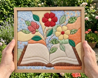 May include: A framed fabric wall art piece featuring a quilted design of an open book with flowers and leaves emerging from the pages. The artwork uses various patterned fabrics in red, yellow, blue, green, and orange, all encased in a light wood frame.