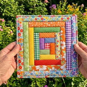 May include: A square fabric quilt block featuring a log cabin pattern constructed from various colorful cotton strips. The design incorporates floral, gingham, and polka dot prints in shades of orange, green, yellow, red, purple, and blue.