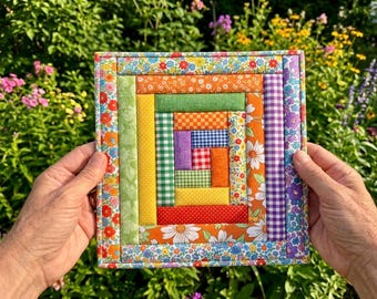 May include: A square fabric quilt block featuring a log cabin pattern with concentric strips of colourful cotton fabric. The design incorporates various floral, gingham, and polka dot prints in shades of orange, yellow, green, blue, red, and purple.