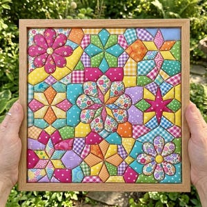 Puede incluir: Un marco de madera cuadrado que contiene un bloque de edredón de tela colorido con un patrón geométrico. El diseño presenta varias formas, incluyendo estrellas y flores en tonos rosa, amarillo, naranja, azul y verde, con estampados de lunares y cuadros.