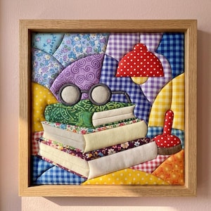 May include: A square fabric wall art piece in a light wood frame featuring a quilted patchwork design. The image depicts a stack of three books topped with round spectacles, a red polka-dot lamp, and a red polka-dot pincushion on a colorful background.