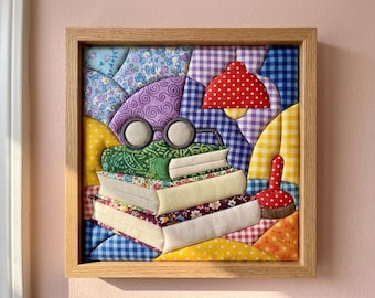 Op de afbeelding: Een vierkant stoffen wandkunstwerk in een lichthouten lijst met een gewatteerd patchworkontwerp. De afbeelding toont een stapel van drie boeken met daarop een ronde bril, een rood lampje met stippen en een rood speldenkussen met stippen.