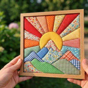 Könnte beinhalten: Ein quadratisches Wandbild aus Stoff in einem hellen Holzrahmen mit einer gesteppten Sonnen- und Berglandschaft. Das Design verwendet verschiedene gemusterte Stoffe in Rot, Orange, Gelb, Blau und Grün, darunter Blumen-, Vichy- und Punktmuster.