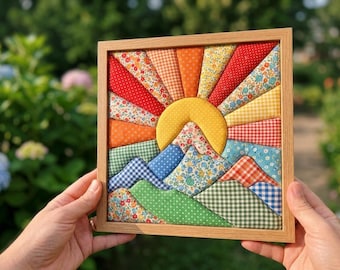 May include: A square fabric wall art piece in a light wood frame featuring a quilted sun and mountain landscape. The design uses various patterned fabrics in red, orange, yellow, blue, and green, including floral, gingham, and polka dot prints.
