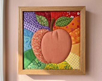 May include: A square fabric wall art piece in a light wood frame featuring a central peach-coloured apple with a brown stem and two green leaves. The background is composed of radiating fabric segments in purple, blue, green, yellow, orange, and red.
