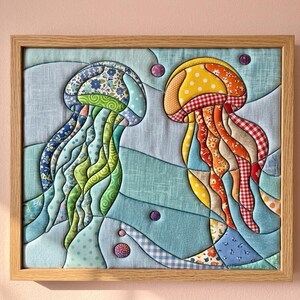 May include: A framed fabric wall art piece featuring two jellyfish appliqu&eacute;s made from various patterned textiles. The left jellyfish uses blue, green, and floral fabrics, while the right one uses yellow, orange, and red gingham and floral prints.