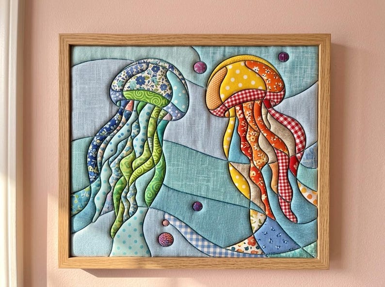 May include: A framed fabric wall art piece featuring two jellyfish appliqu&eacute;s made from various patterned textiles. The left jellyfish uses blue, green, and floral fabrics, while the right one uses yellow, orange, and red gingham and floral prints.