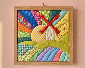 May include: A square fabric wall art piece in a light wood frame featuring a quilted landscape scene. The design includes a central windmill with red patterned blades, a yellow striped sun, and various colourful fabric patches in floral, polka dot, and gingham patterns representing rolling hills and a bright sky.