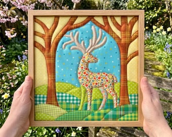 May include: A square fabric wall art piece in a light wood frame featuring a quilted deer with a floral pattern and large antlers. The background depicts a blue sky with small stars, orange plaid trees, and green hills with various patterns.