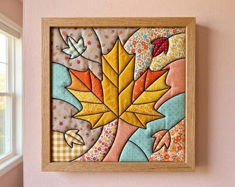May include: A square wall art piece featuring a central yellow maple leaf surrounded by smaller leaves in a quilted fabric design. The artwork is encased in a light-coloured wooden frame and displays various patterns including floral, gingham, and solid colours.