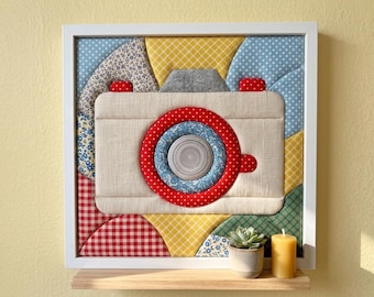 May include: A square framed fabric wall art piece featuring a quilted camera design. The camera is beige with a red polka dot lens ring and blue floral center. The background consists of various colorful patterned fabric patches in yellow, blue, and red.