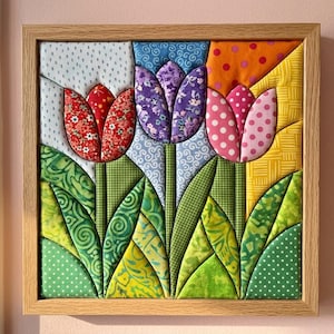 May include: A square fabric wall art piece in a light wood frame featuring three dimensional quilted tulips. The flowers are red, purple, and pink with various patterns, set against a background of blue, orange, and yellow fabric patches.